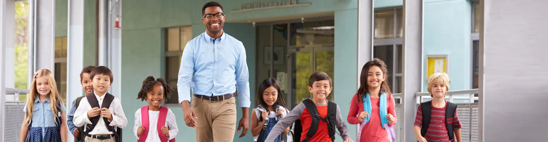 A man in a blue shirt walks forward with a group of children wearing backpacks outside a building.