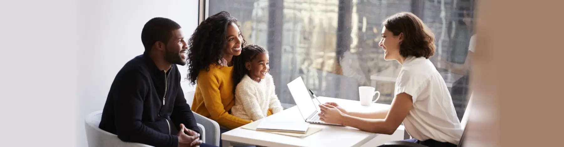 A woman sits at a white table with a laptop facing a man, a woman, and a young girl who are seated together and smiling.