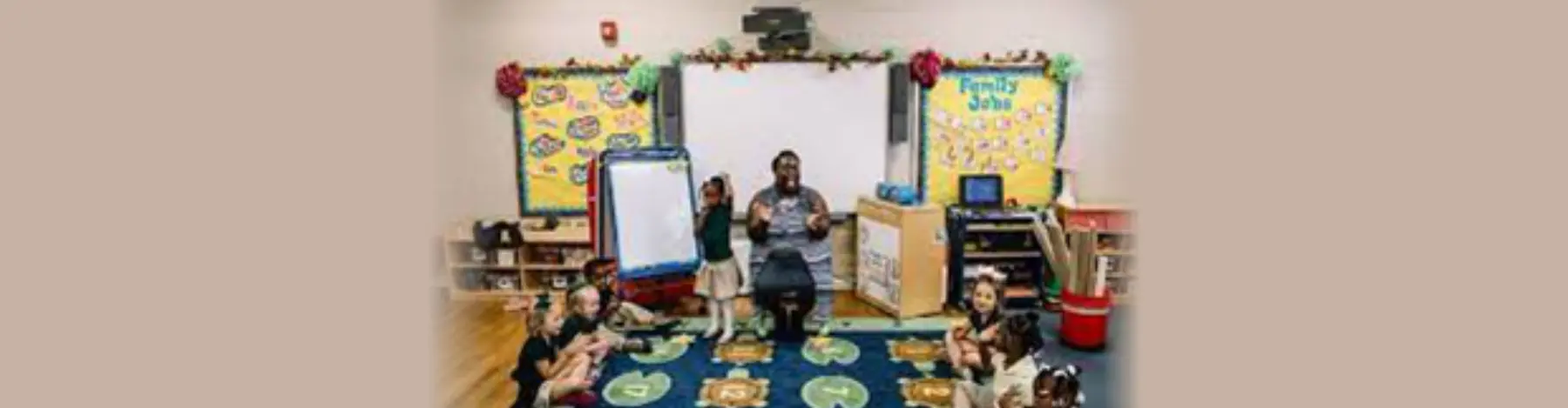 A teacher sits on a colorful rug facing several children seated around her in a classroom with educational posters on the walls.