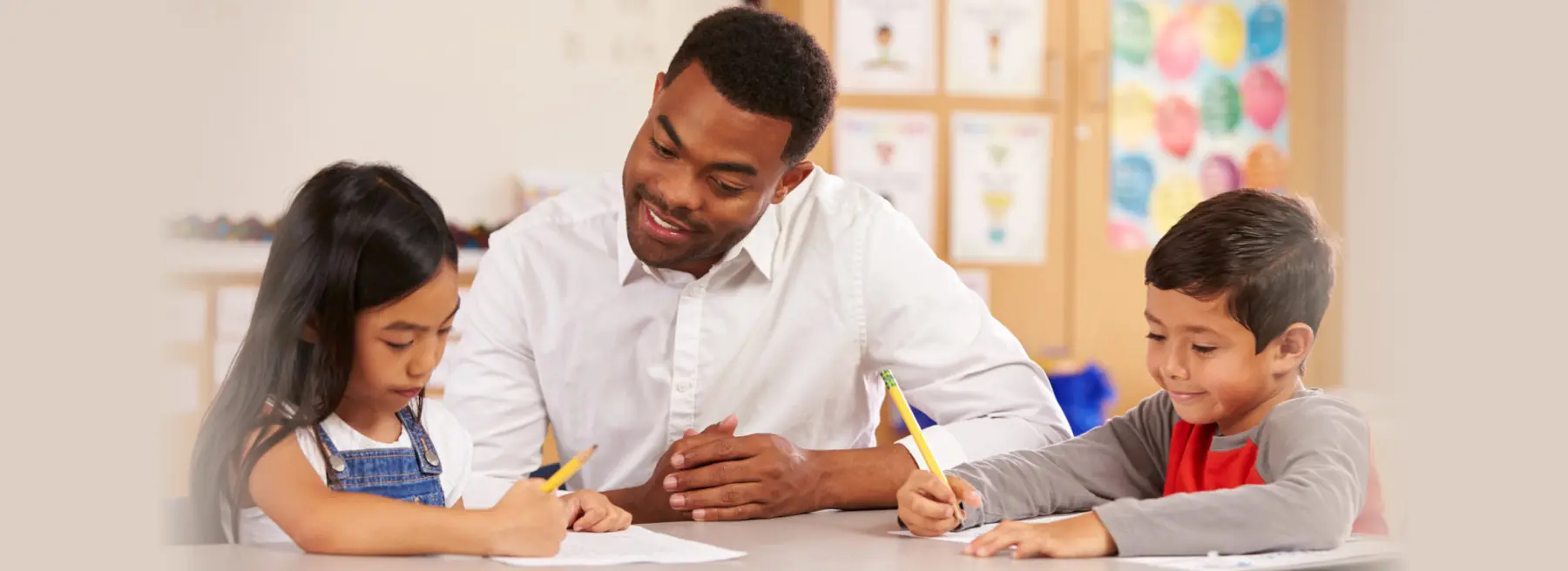 A man in a white shirt sits between two children at a table, watching as they write on papers with yellow pencils.