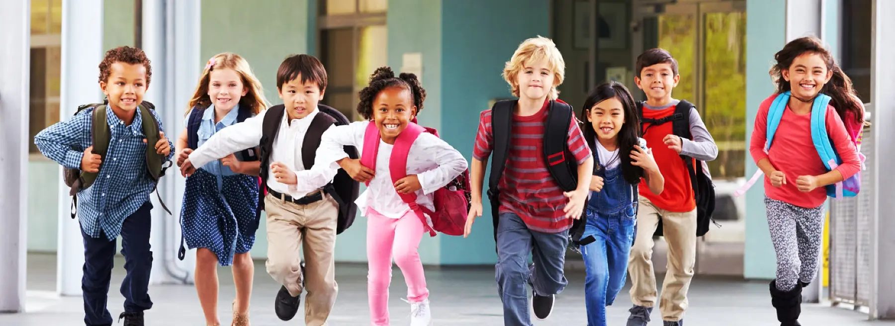 Eight children wearing backpacks run toward the camera along a paved path beside a building with large windows.