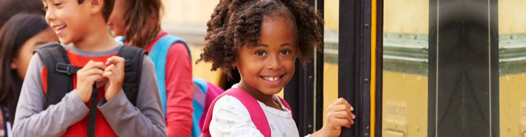 Children wearing backpacks stand beside a yellow school bus, with a smiling girl in the foreground holding the bus door.