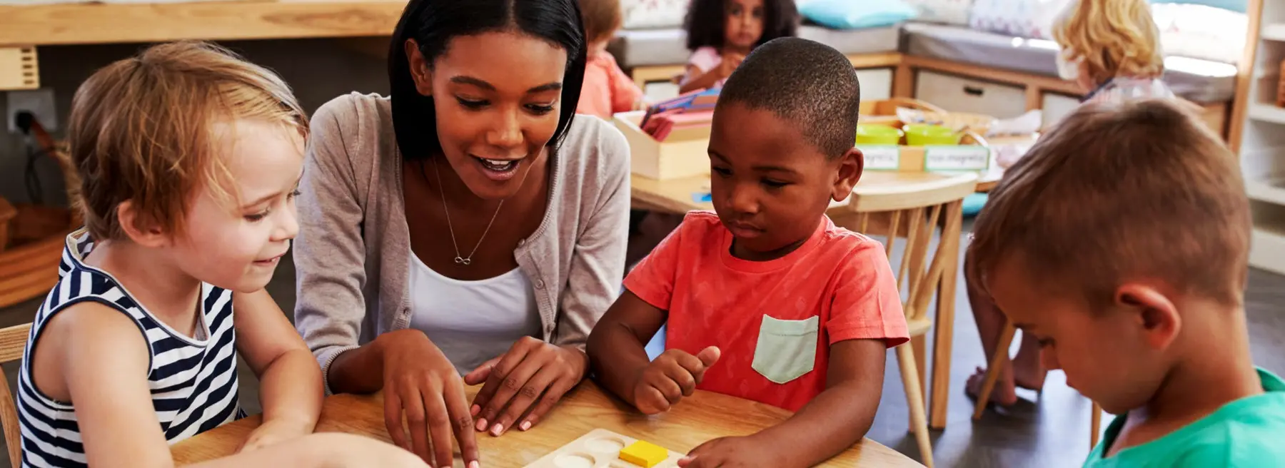 A woman sits at a low table with three children, pointing at a small yellow object while the children watch and reach toward it.