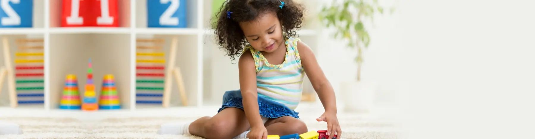 A young girl sits on a rug and plays with colorful toy building blocks in front of a white shelf holding bins and stacked rings.