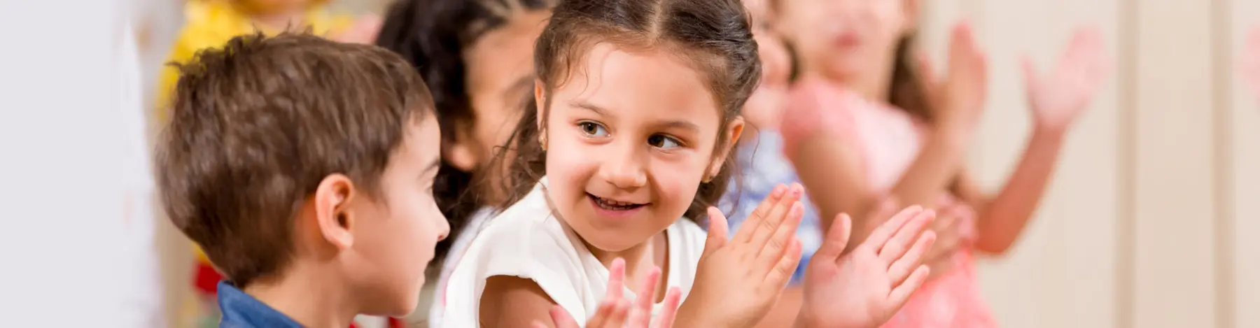Several young children sit in a row clapping their hands, with a girl in a white top smiling in the foreground and a boy in a blue shirt beside her.