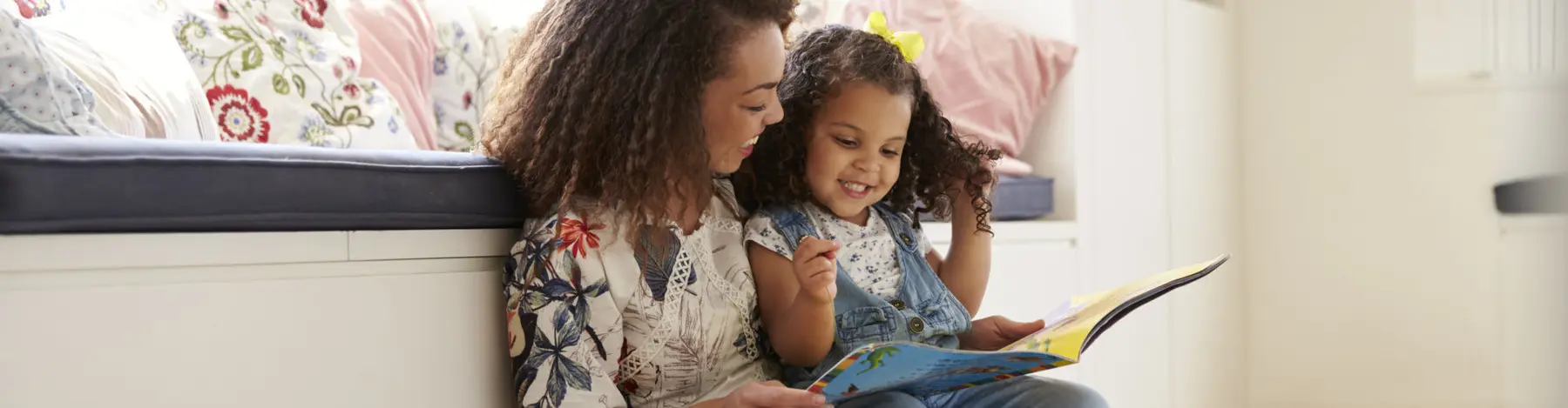 A woman and a young girl sit together on a bench reading a colorful book.
