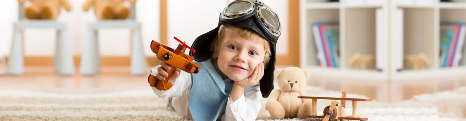A young child wearing aviator goggles and a pilot hat lies on a rug, holding a wooden toy airplane and resting their chin on their hand next to a teddy bear.