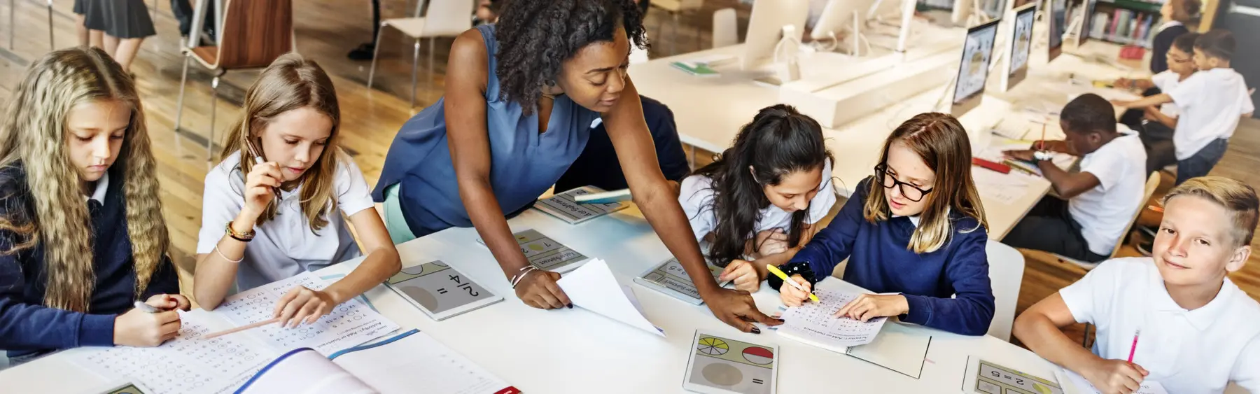A woman leans over a table to help several children working in notebooks, while other children use laptops at desks in the background.