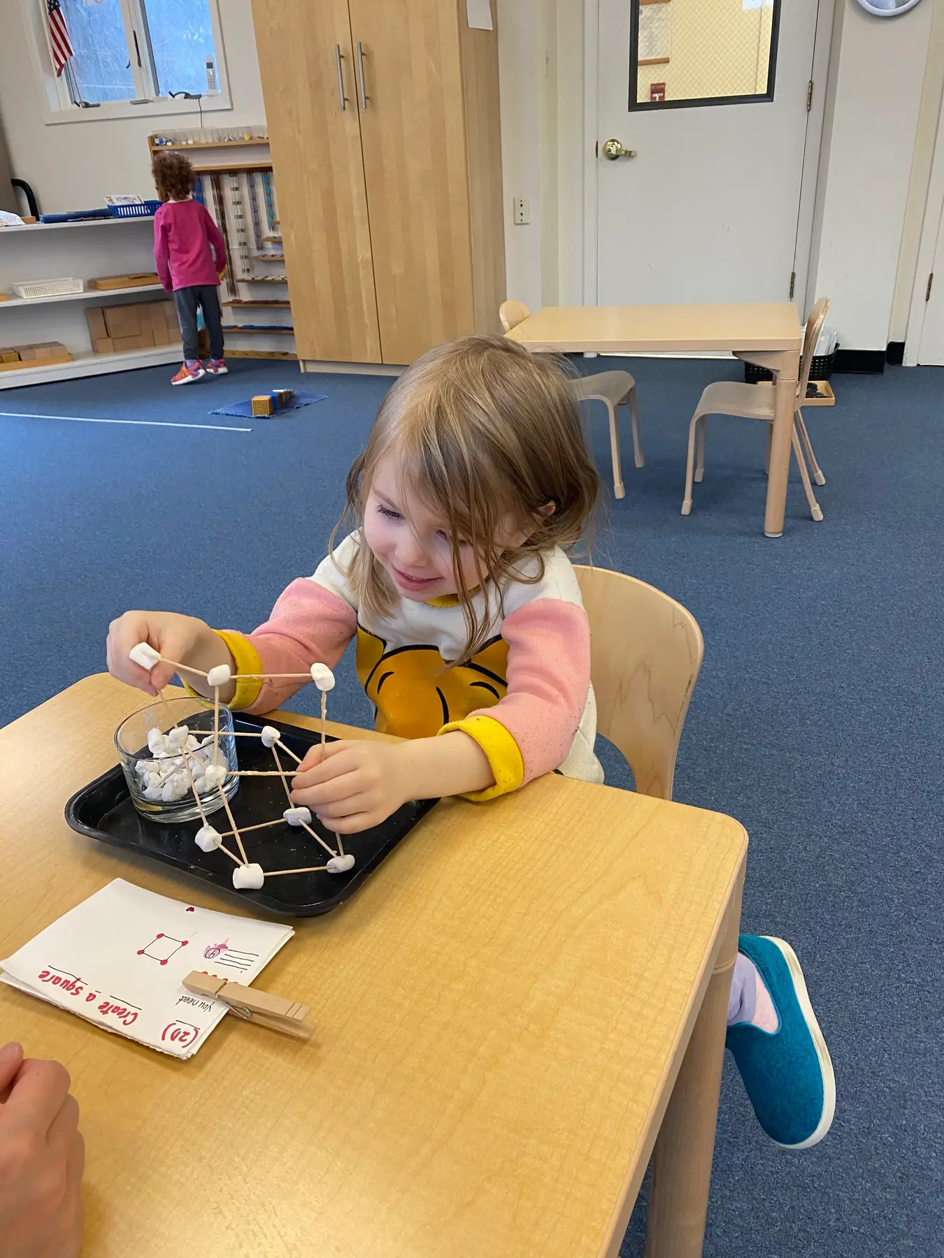 A young girl sits at a table building a geometric structure with toothpicks and marshmallows while another child stands near shelves in the background.