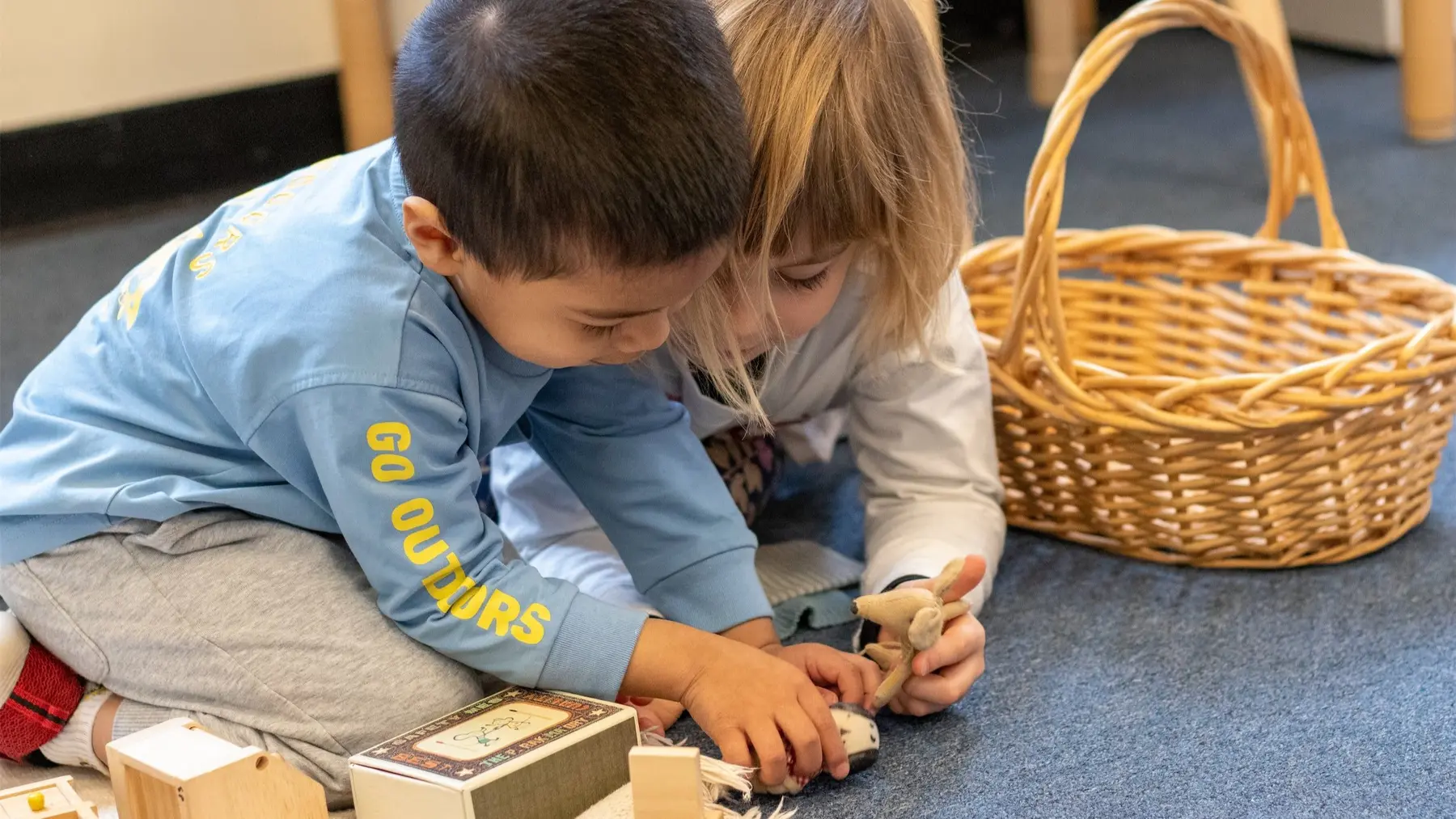 Two young children sit on a carpeted floor, leaning over a small wooden toy and blocks, with a woven basket nearby.