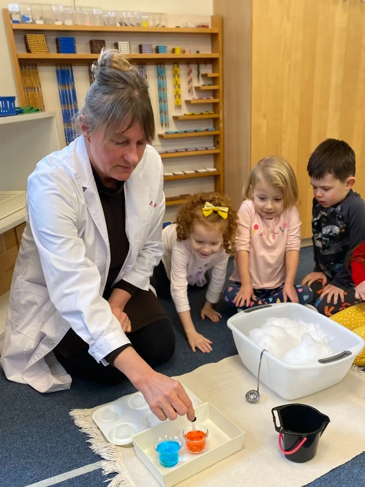 A woman in a white coat kneels on a rug and points at two small cups of colored liquid while three children sit and watch beside a bin of white material.