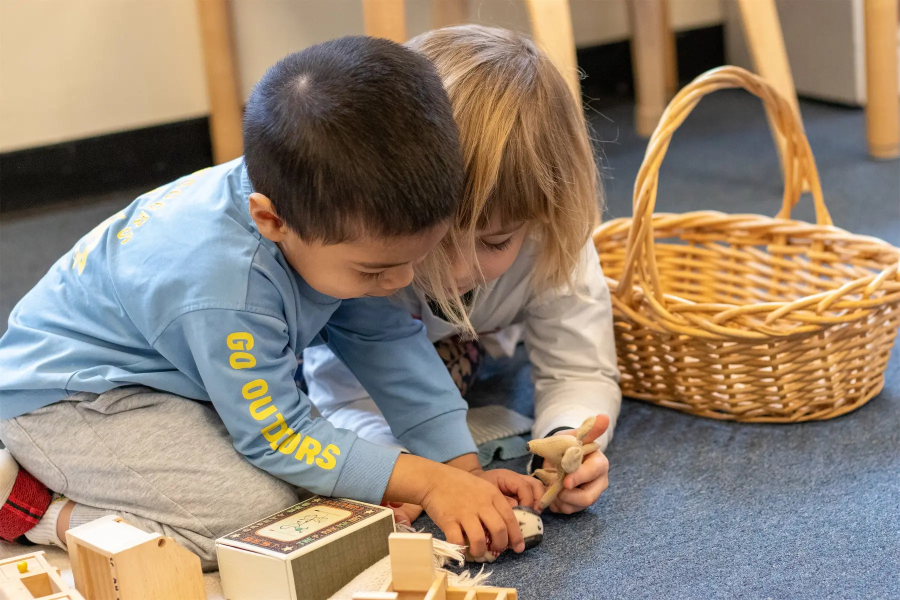 Two children sit on a carpeted floor, looking down as they play with small wooden toys and blocks next to a woven basket.
