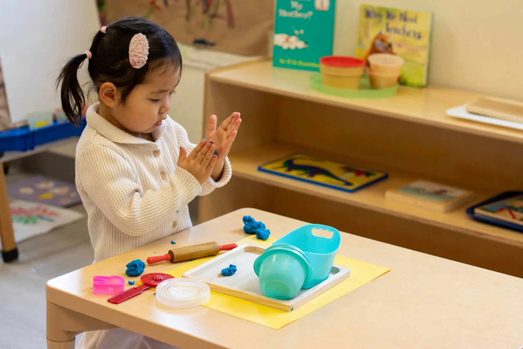 A young girl in a white sweater stands at a table with her hands clasped together over a tray of blue play dough and plastic tools.
