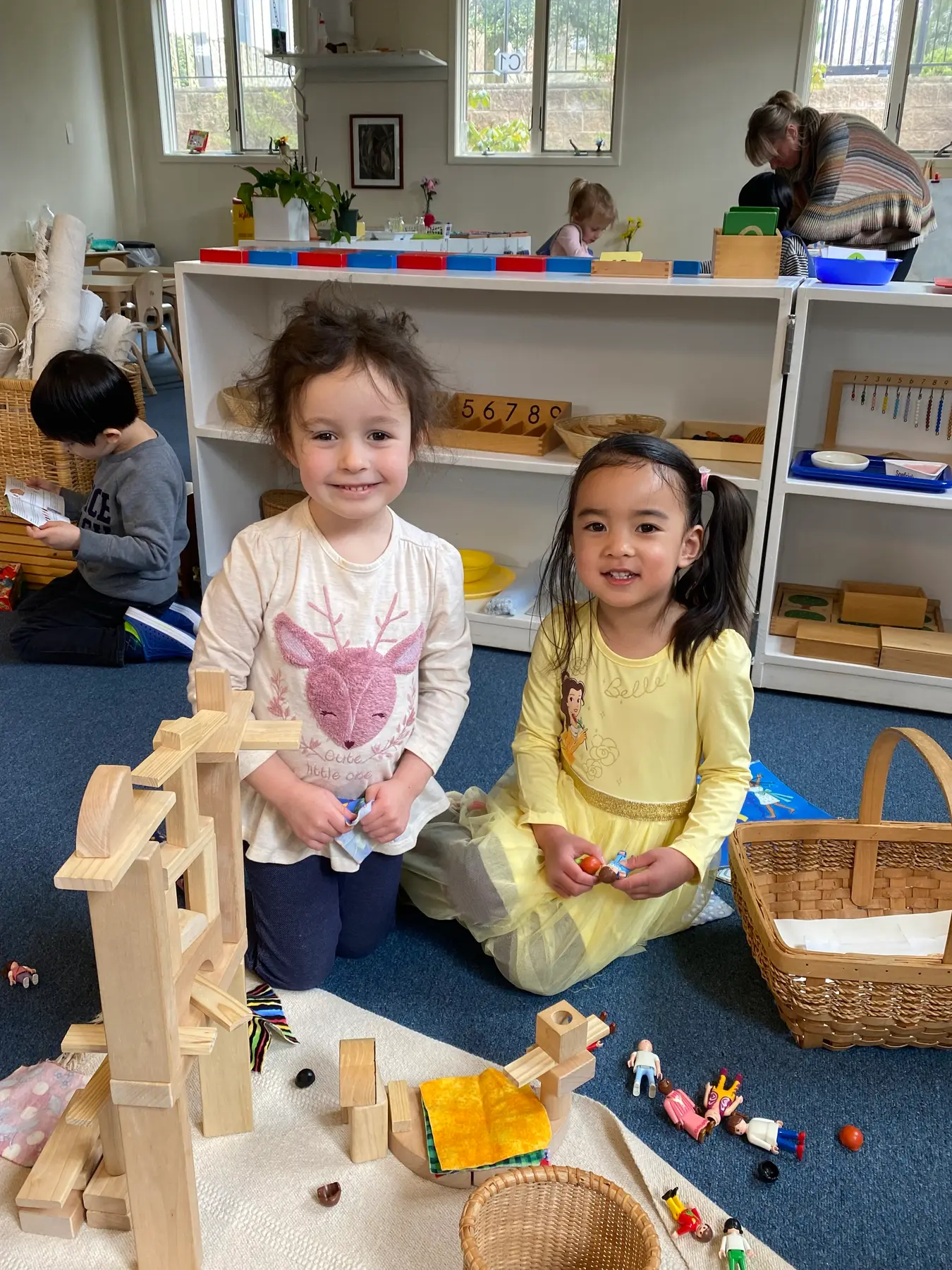 Two girls kneeling on a blue carpet smile beside a wooden block structure while other children and an adult work at shelves in the background.