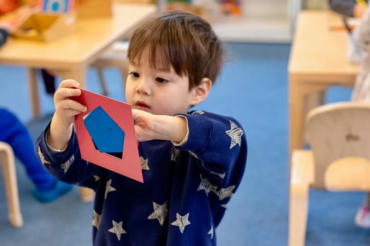 A young boy in a blue star-patterned sweater holds up a red paper cutout with a blue pentagon shape inside while standing in a classroom.