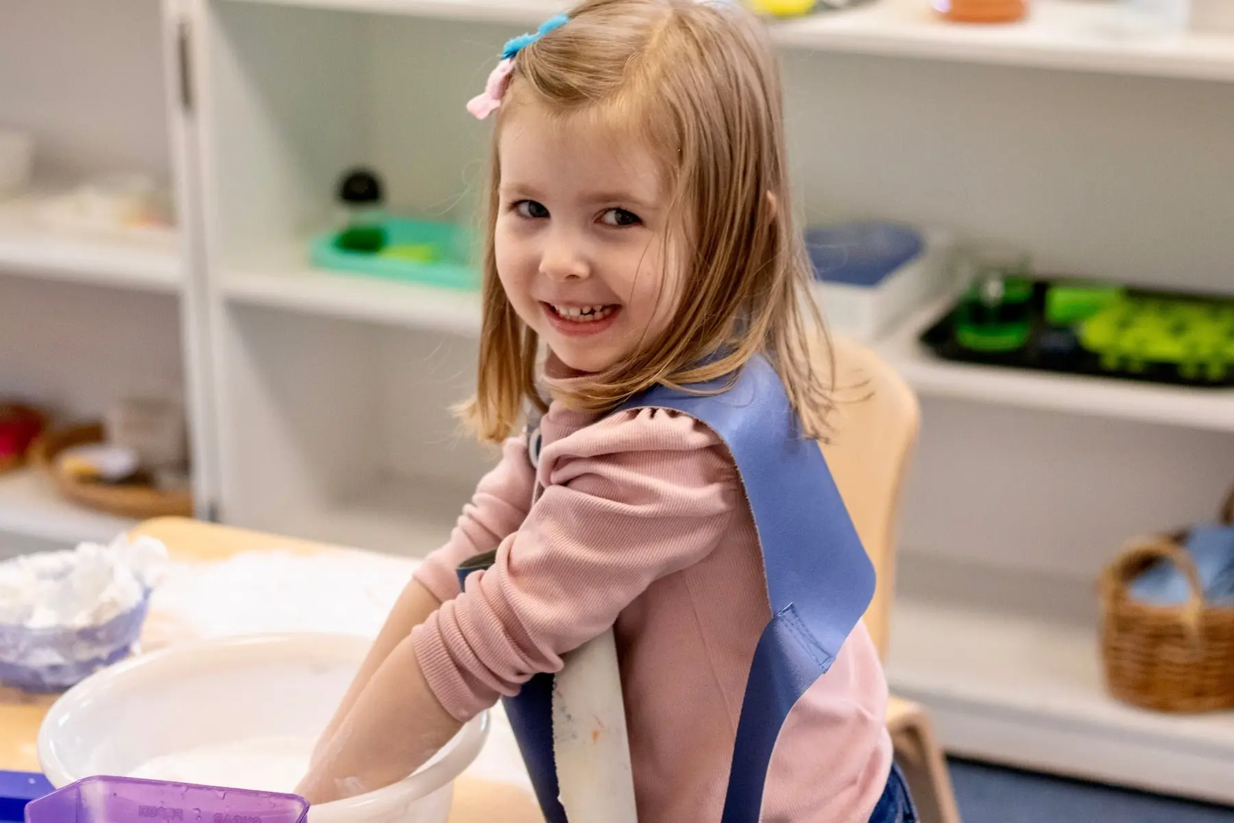 A young girl in a pink shirt and blue apron smiles while standing at a table with a white bin and a purple container.