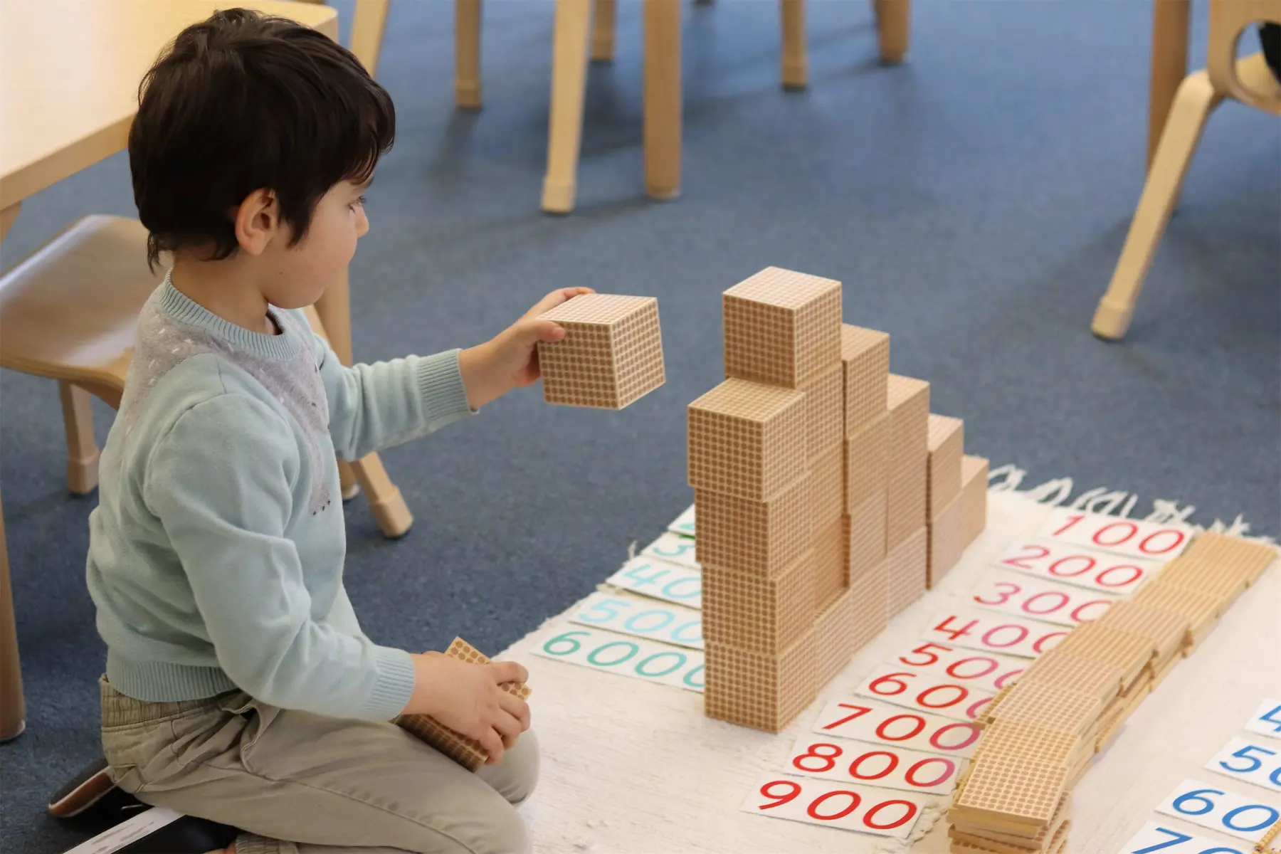 A young boy sits on a rug and holds a small wooden cube above a stack of base-ten blocks arranged in a staircase beside number cards.