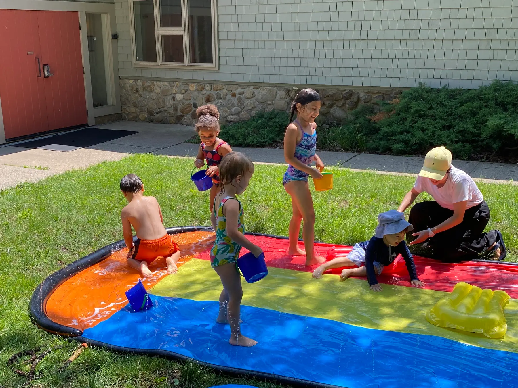 Five children in swimsuits play on a colorful water mat while an adult in a cap kneels beside them on the grass.