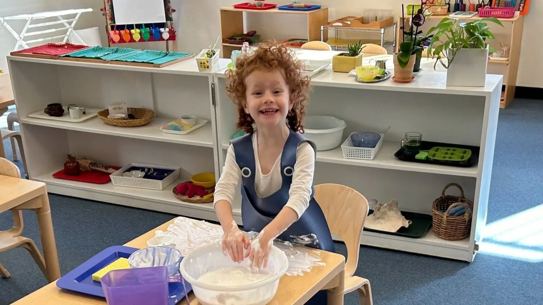 A smiling child with curly hair stands at a table wearing a blue apron, pressing hands into a white bowl of flour mixture.