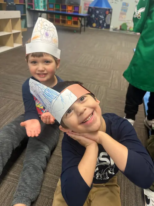 Two boys sit on a carpeted floor wearing paper headbands with drawings, one with hands under his chin and the other with a hand raised, in a room with shelves and toys.