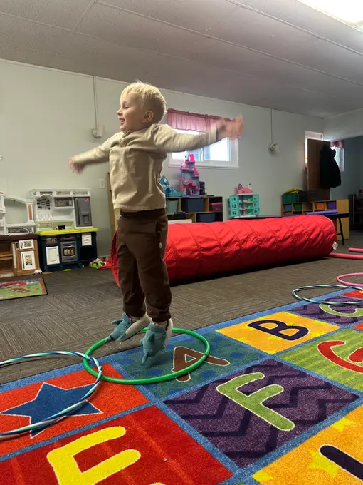 A young child stands with arms outstretched inside a green hula hoop on a colorful alphabet rug in a room with toys and a red mat.