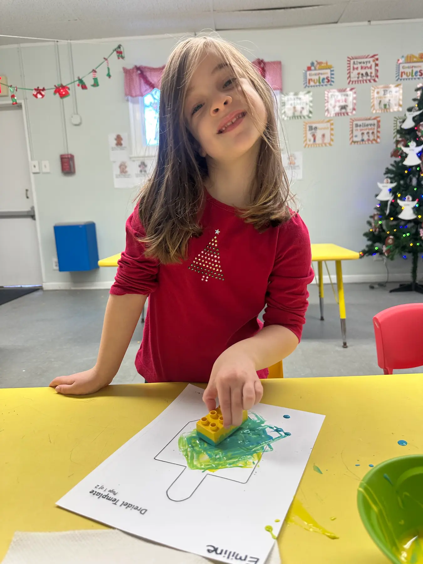 A girl in a red shirt with a tree design stands at a yellow table pressing a yellow building block into green paint on a paper template.