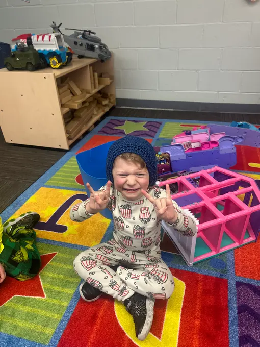 A child wearing a blue knit hat and patterned pajamas sits cross-legged on a colorful rug, holding up two fingers on each hand while surrounded by toys and shelving.
