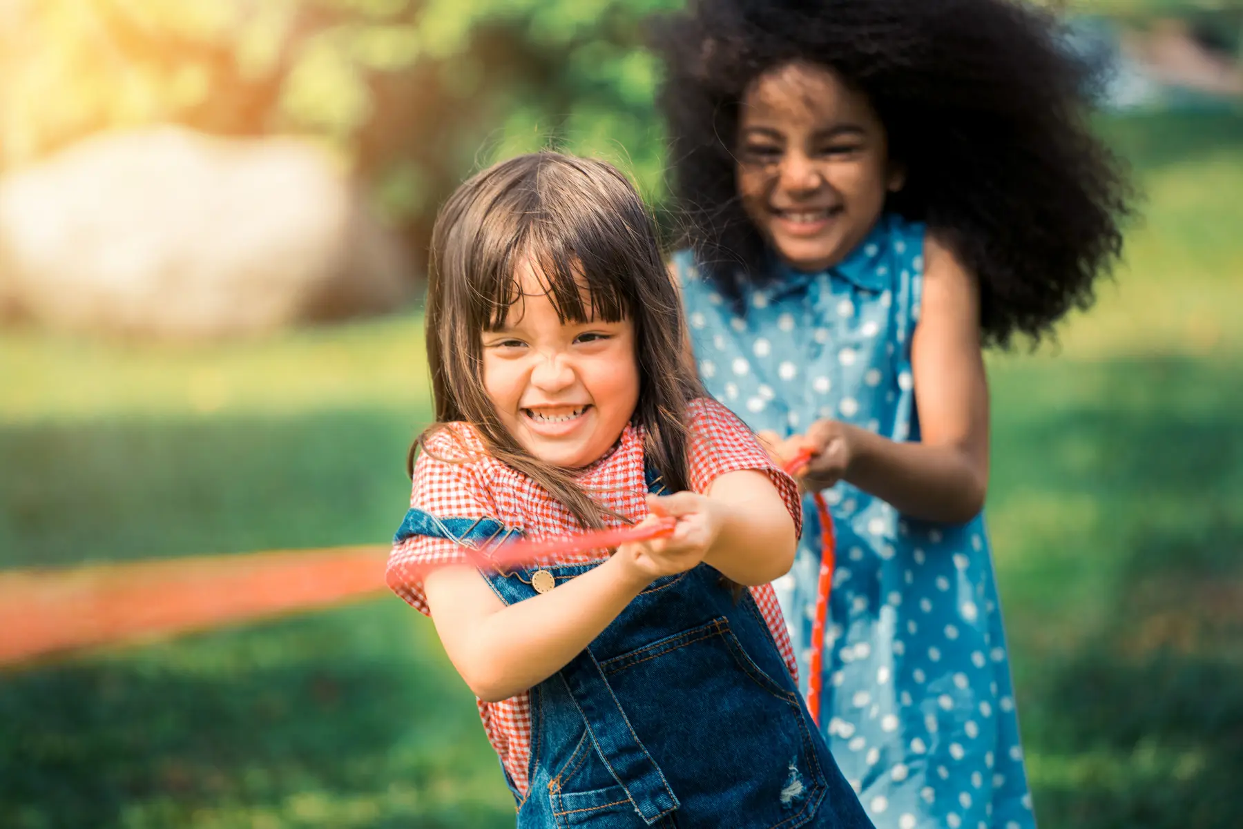 Two smiling girls pull a red rope in a game of tug-of-war on a grassy lawn.