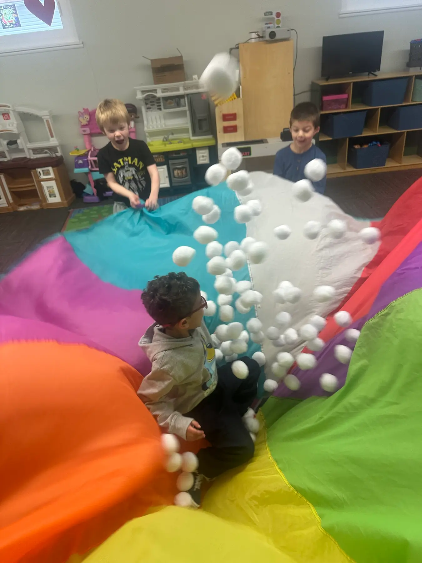 Three children play with a large multicolored parachute, tossing white balls into the air in a classroom.