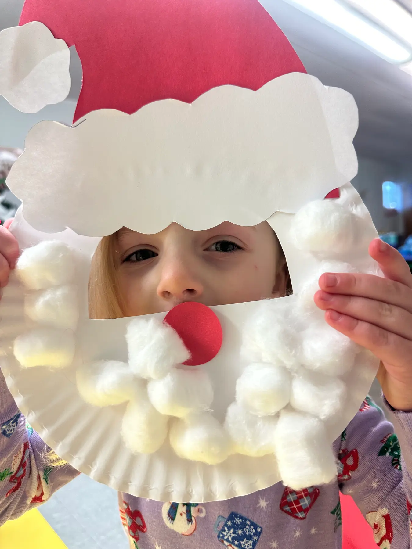 A child holds a paper plate Santa Claus craft with cotton ball beard and red paper hat up to their face.