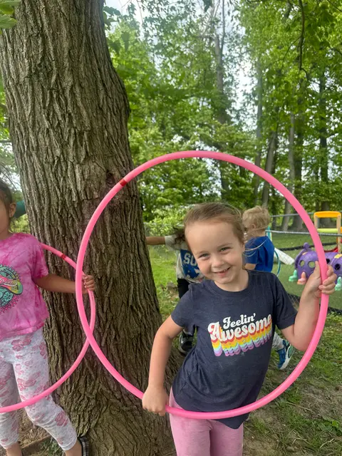 Two girls in the foreground hold pink hula hoops near a large tree while other children play in the grassy background.