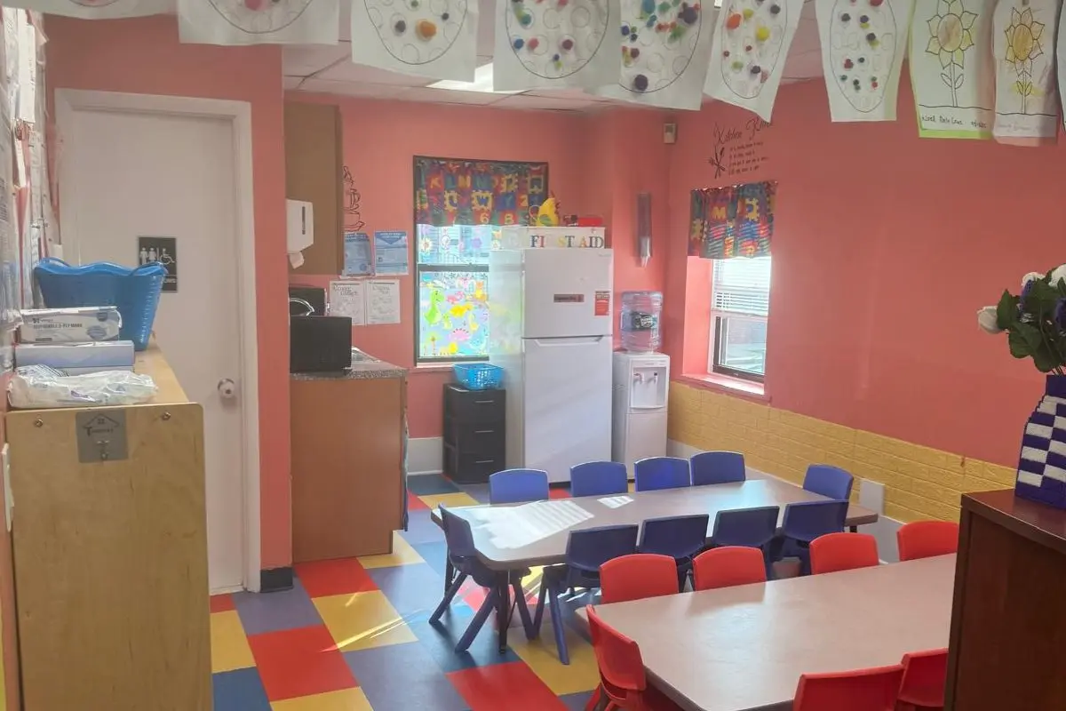 An empty childcare classroom with small tables, blue and red chairs, a refrigerator, and hanging artwork on a pink wall.
