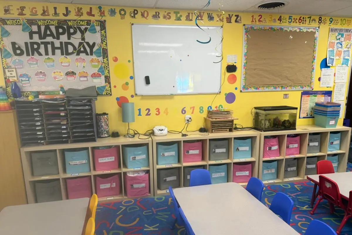 A preschool classroom with yellow walls, alphabet and number decorations, a birthday banner, a whiteboard, storage cubbies with colorful bins, tables with chairs, and a fish tank.