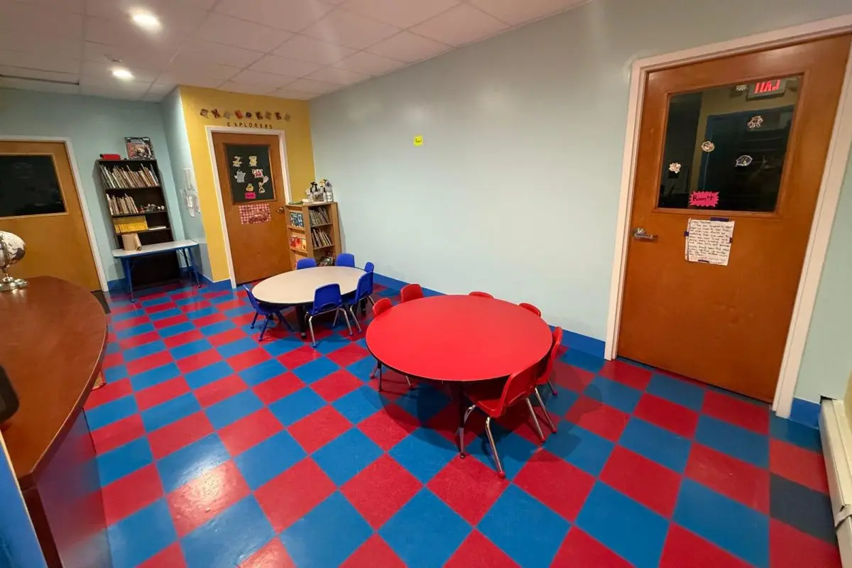 An empty classroom with a red and blue checkered floor, two round tables with small chairs, wooden doors, and bookshelves against light blue and yellow walls.