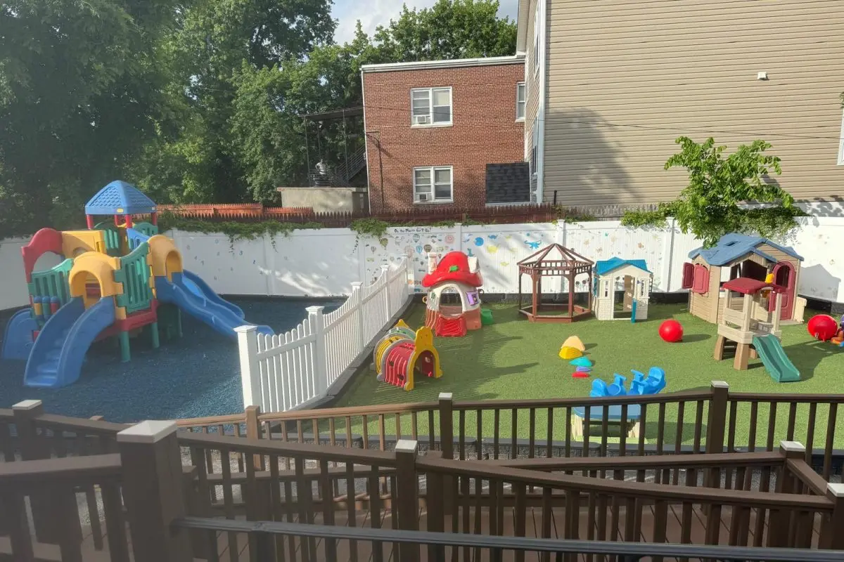 A fenced outdoor play area with a large multicolored play structure on blue surfacing and a smaller playhouse area on green surfacing, surrounded by white fencing and buildings.
