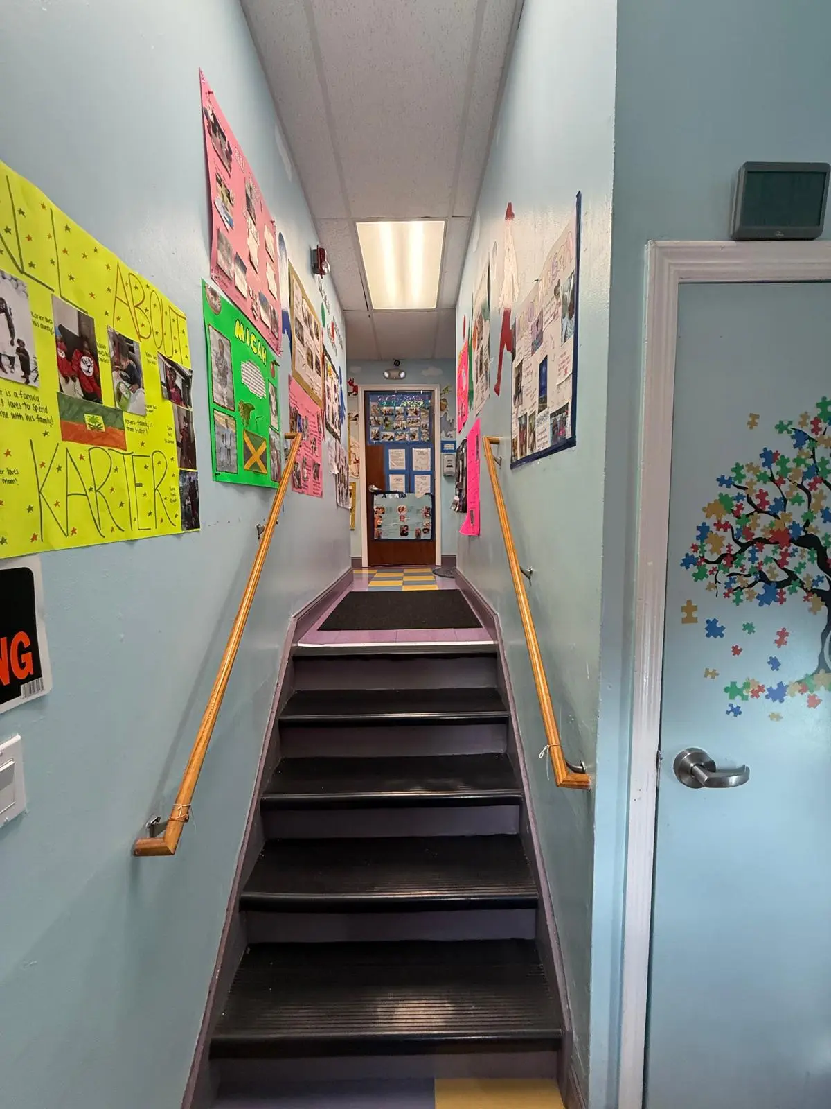A stairwell with blue walls, wooden handrails, and dark steps leads up to a closed door decorated with children's artwork and posters.