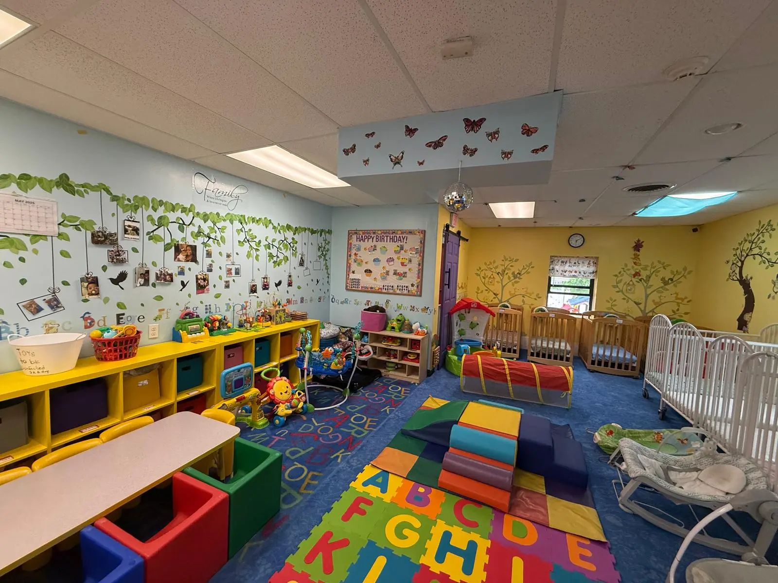 A childcare room with alphabet rugs, yellow storage shelves, cribs, and a wall decorated with leaves and hanging photos.