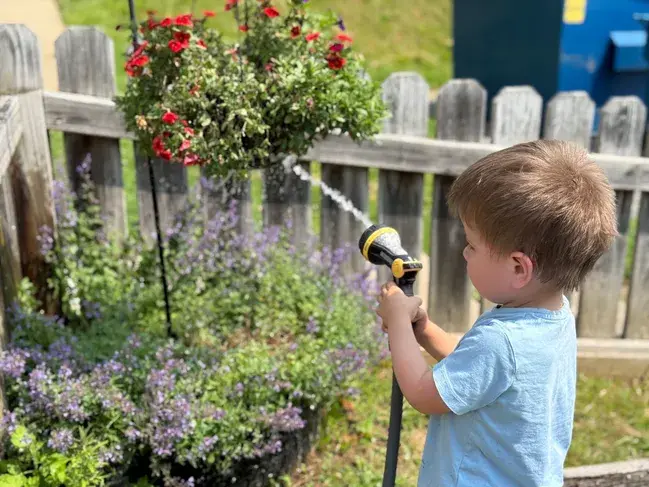 A young child holding a garden hose nozzle watering plants near a wooden fence and hanging flower basket.