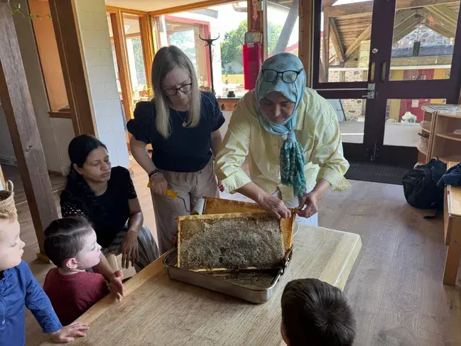 A woman in a headscarf holds a honeycomb frame on a table while several adults and children observe.
