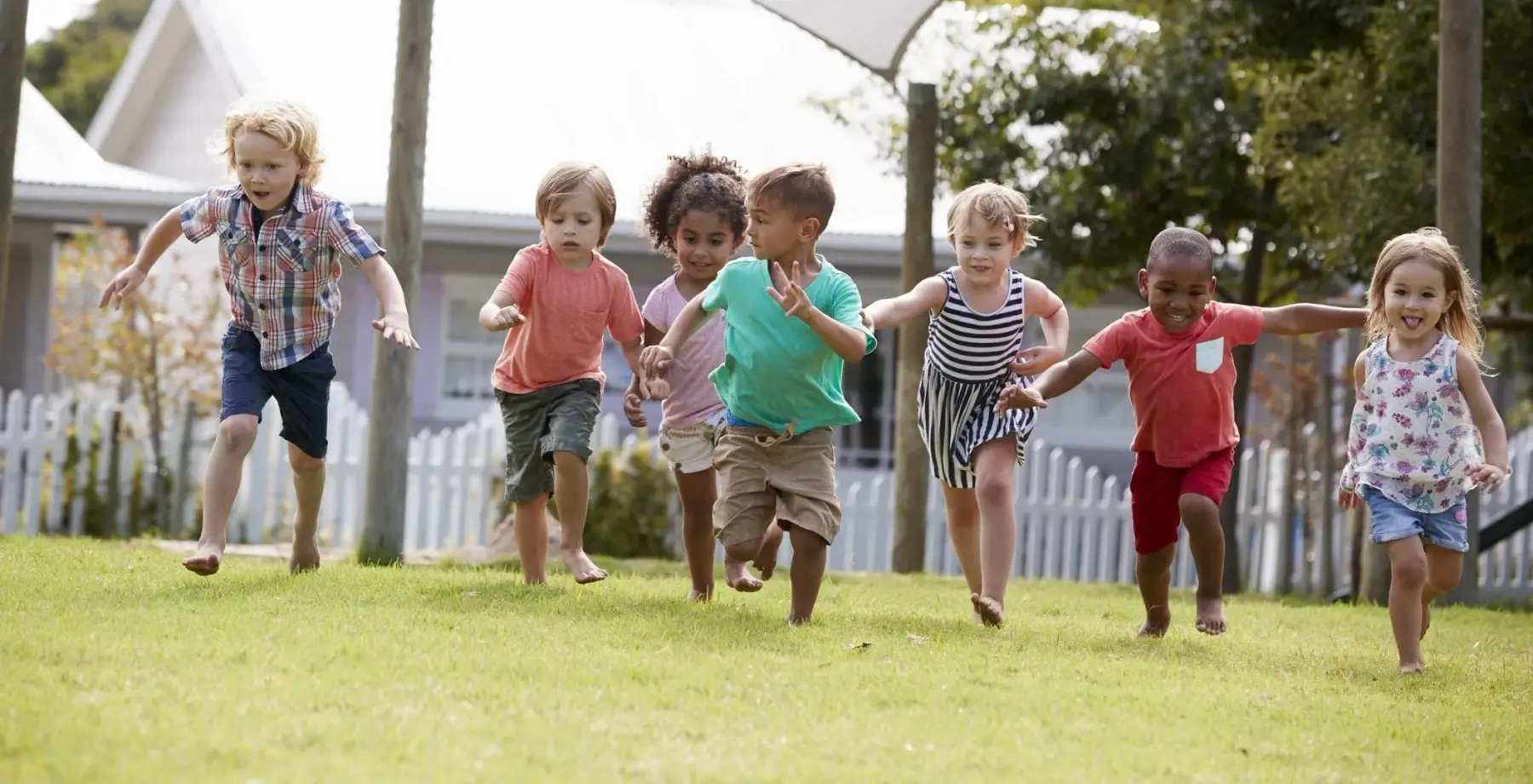 Eight preschool-aged children running together on a grassy lawn with houses and trees in the background.