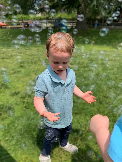 A toddler in a blue polo shirt and dark pants stands on grass with bubbles floating around, while an adult's hand is visible nearby.