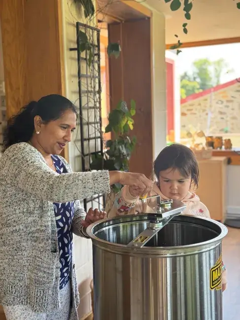 An adult and a toddler interact with a large stainless steel container in an indoor setting.