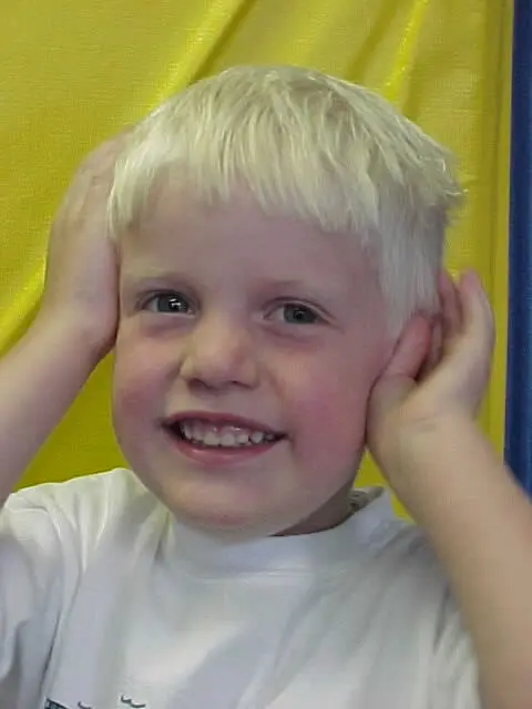 A young child with short blond hair and a white shirt smiles with hands placed on either side of their head against a yellow background.