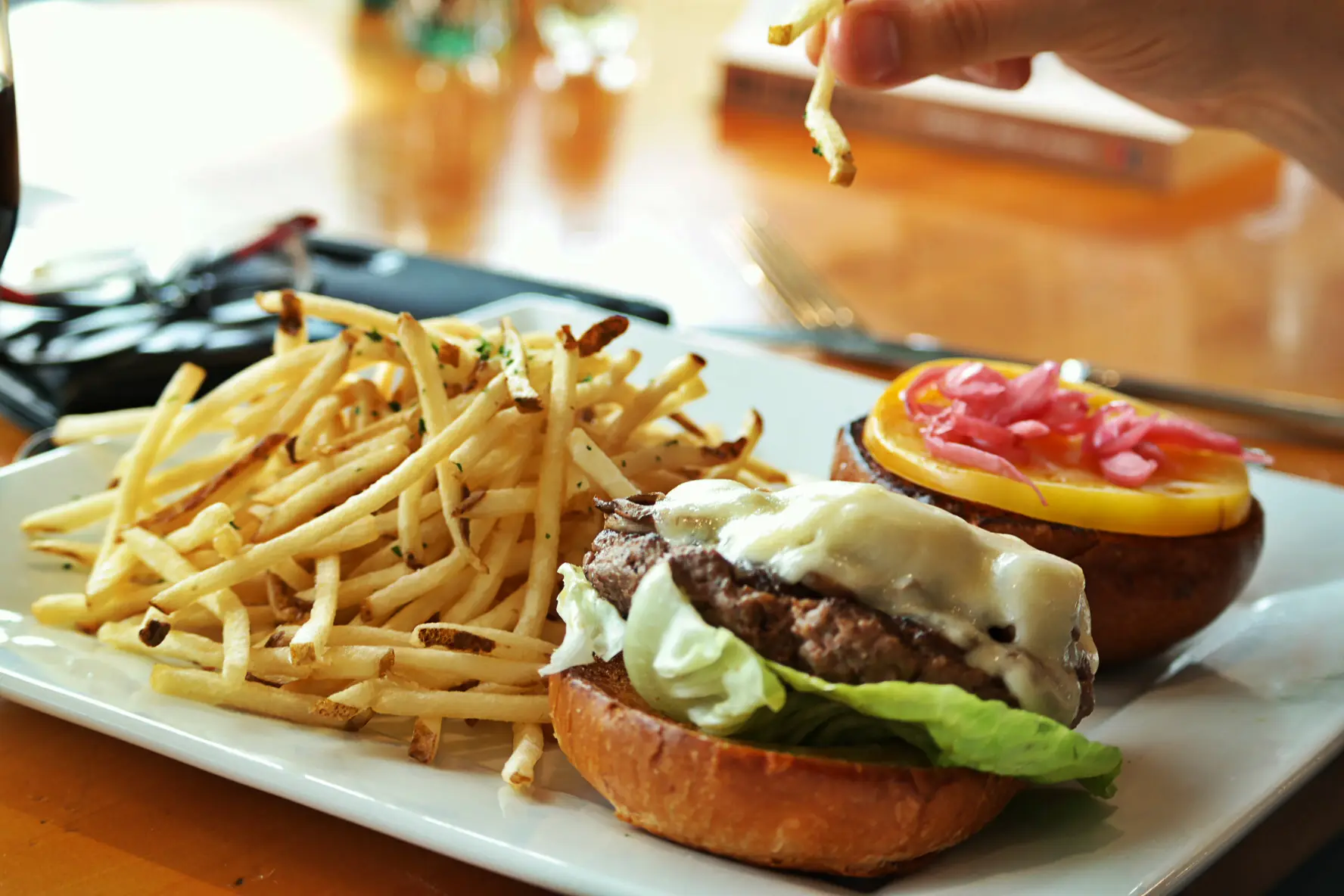 A hand holding a fry above a plate with a burger and fries.