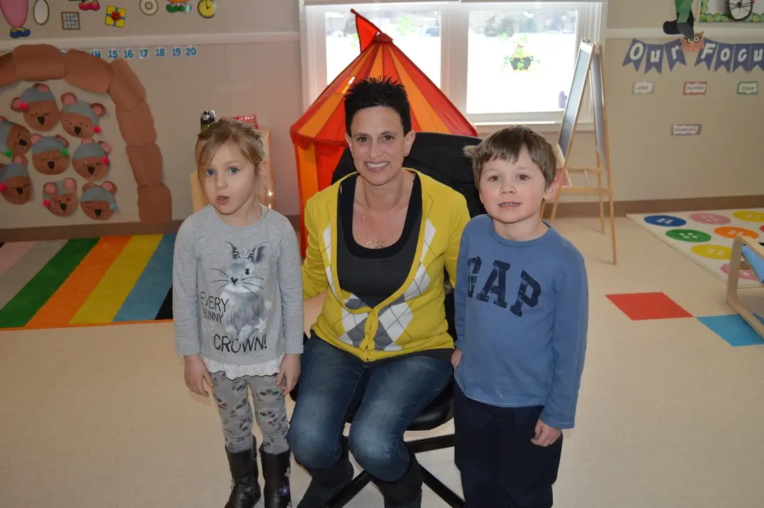 A woman and two children stand in a classroom with colorful rugs, a play tent, and educational wall decorations.