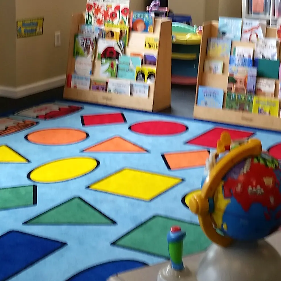 A blue patterned rug with colorful geometric shapes sits in front of wooden bookshelves filled with children's books, with a toy globe in the foreground.