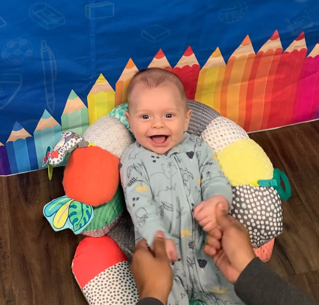 A smiling baby in a patterned sleeper sits in a colorful support pillow while two adult hands hold the baby's hands in front of a blue backdrop with pencil decorations.