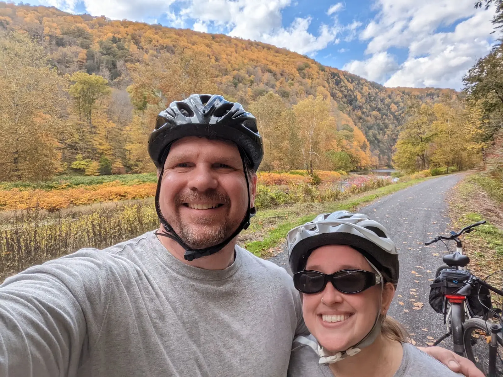 A man and a woman wearing bicycle helmets smile while taking a selfie on a paved path beside a river with autumn trees on a hillside.