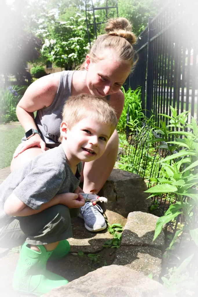 A child crouching on stone steps holds a small toy while an adult crouches behind him in a garden area with plants and a fence.