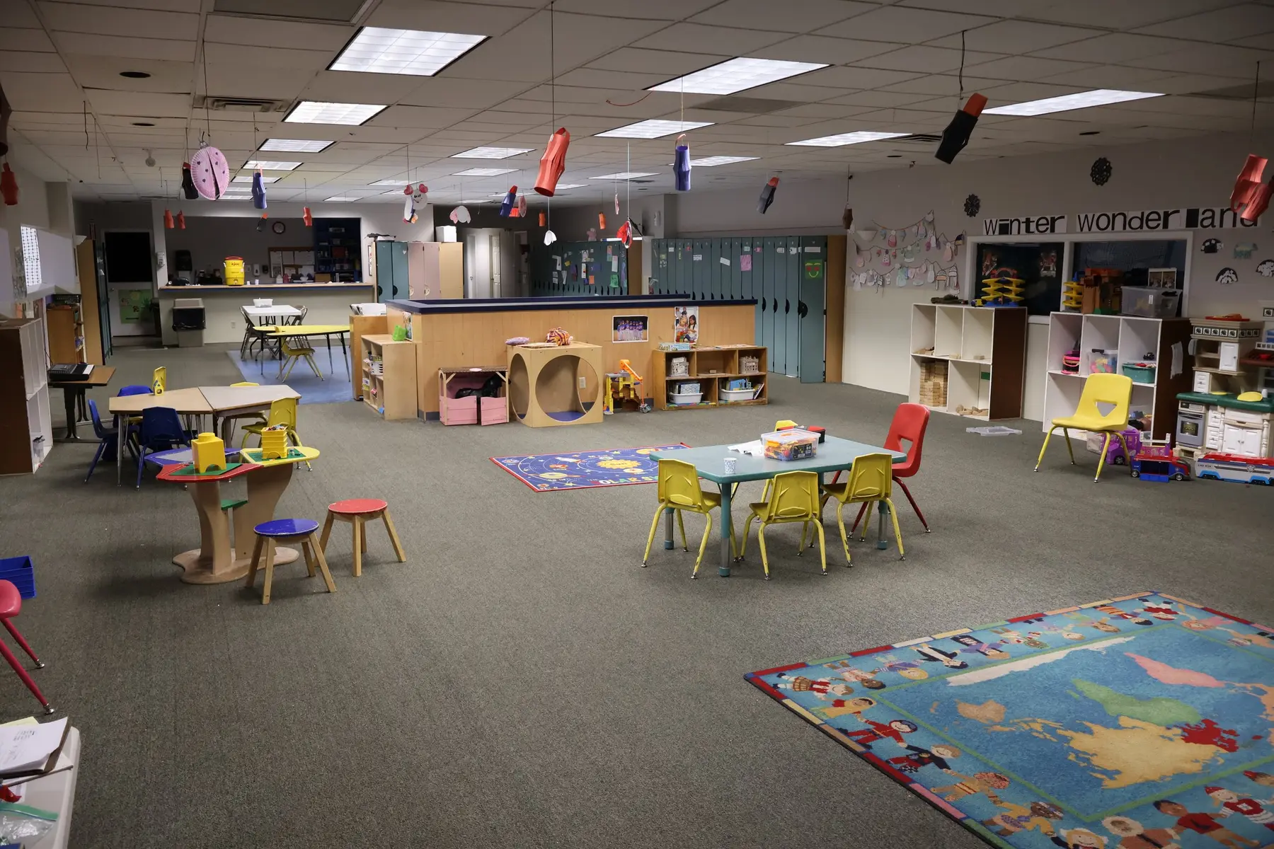 An empty preschool classroom with child-sized tables, colorful chairs, storage shelves, wall cubbies, and two area rugs on a carpeted floor.